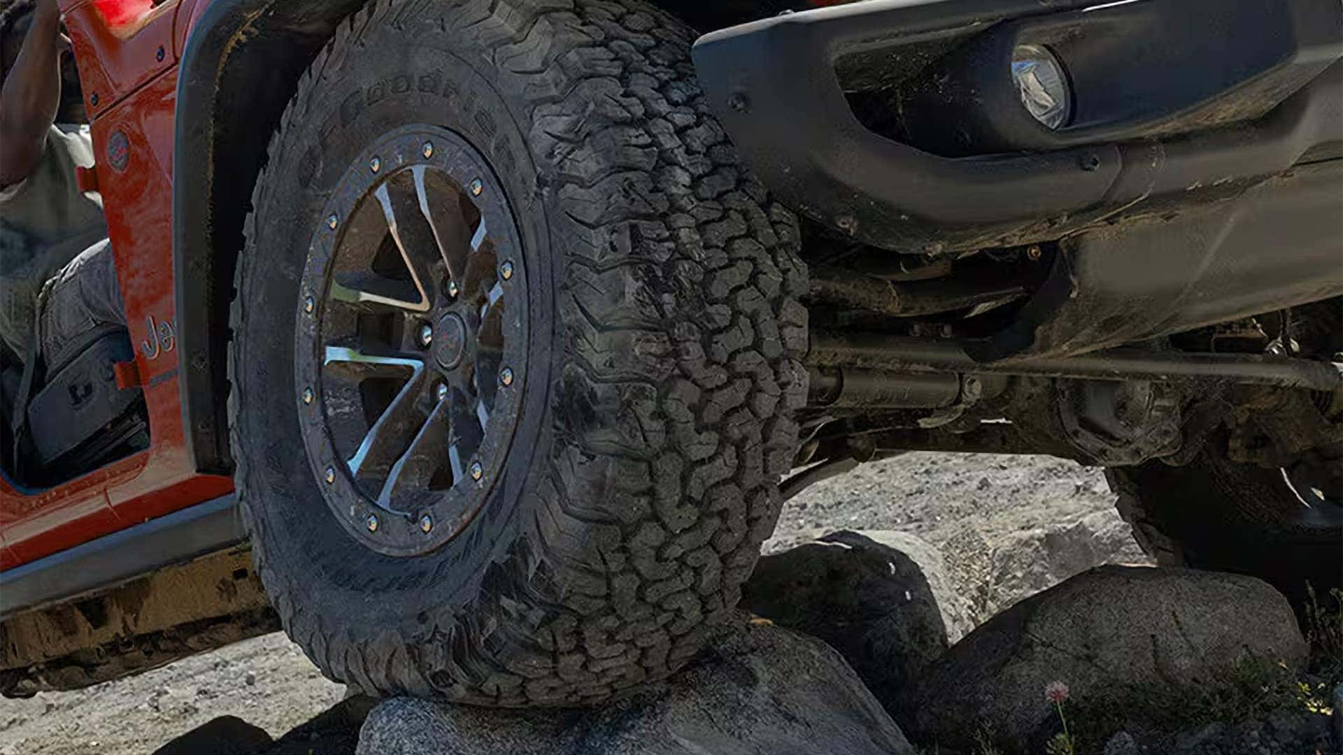 Close-up of a Jeep Wrangler all-terrain tire and alloy wheel climbing rocks, showing rugged tread and suspension.