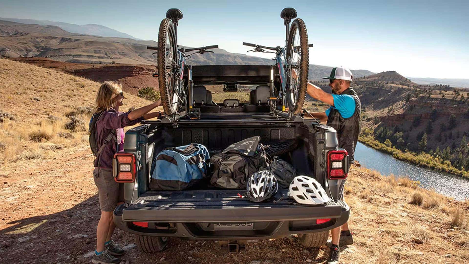 2026 Jeep Gladiator truck bed loaded with outdoor gear as two cyclists secure bikes on a rack near a canyon overlook
