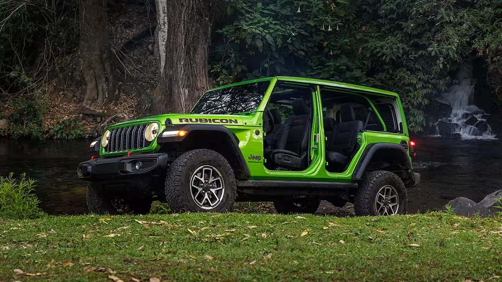 Green 2026 Jeep Wrangler Rubicon with doors removed, parked by a wooded creek with waterfall in the background.