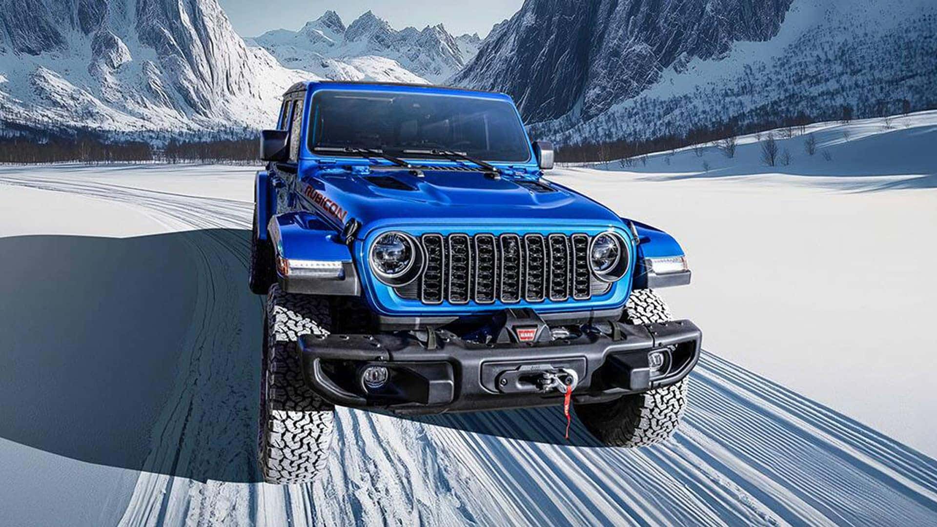 Blue 2026 Jeep Gladiator Rubicon on a snowy mountain road showing seven-slot grille, off-road bumper, and winch.