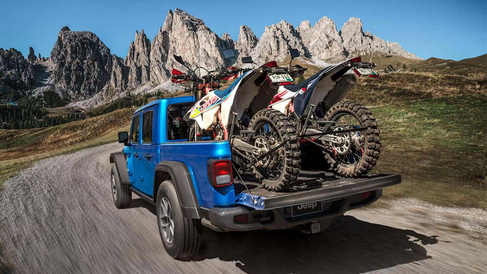 Blue 2026 Jeep Gladiator hauling two dirt bikes in the bed with tailgate down on a mountain road