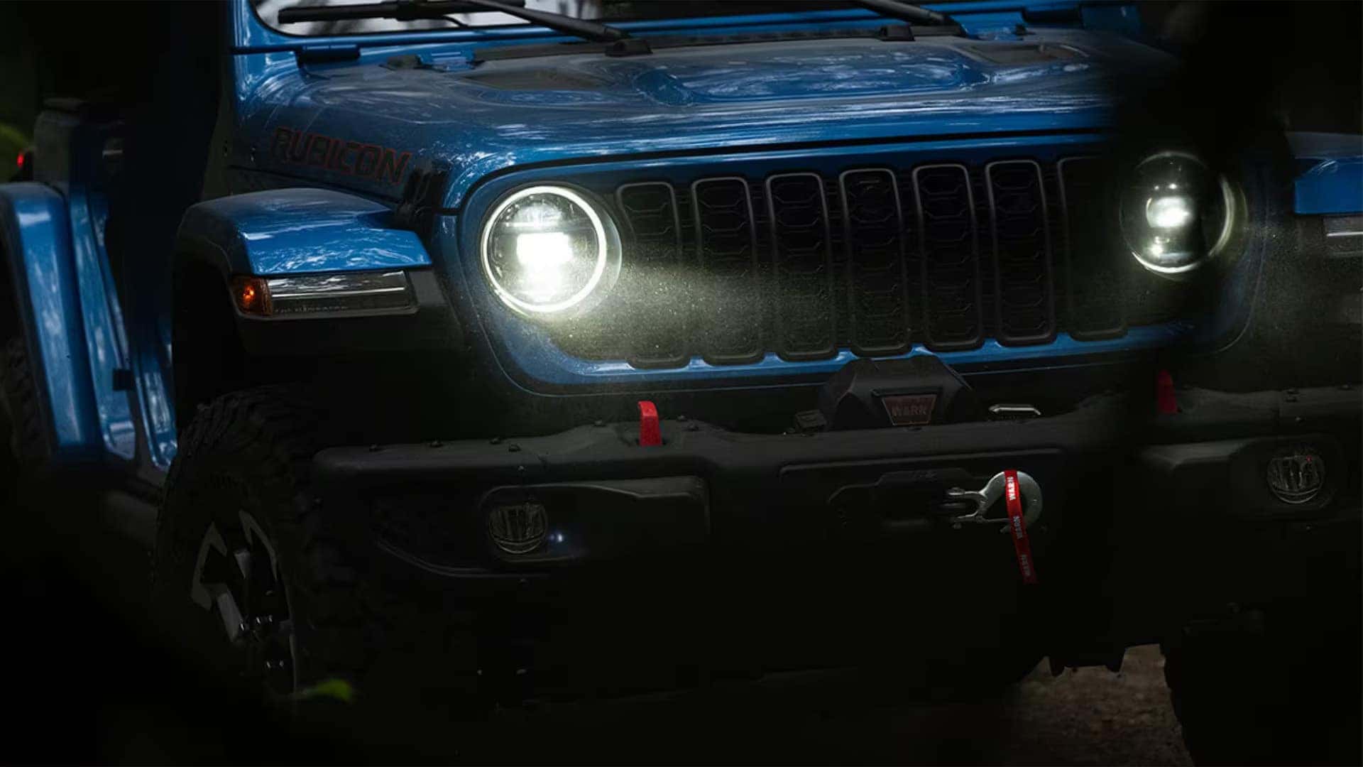 Front view of a blue Jeep Wrangler Rubicon at night with headlights on, seven-slot grille, and winch-ready bumper.