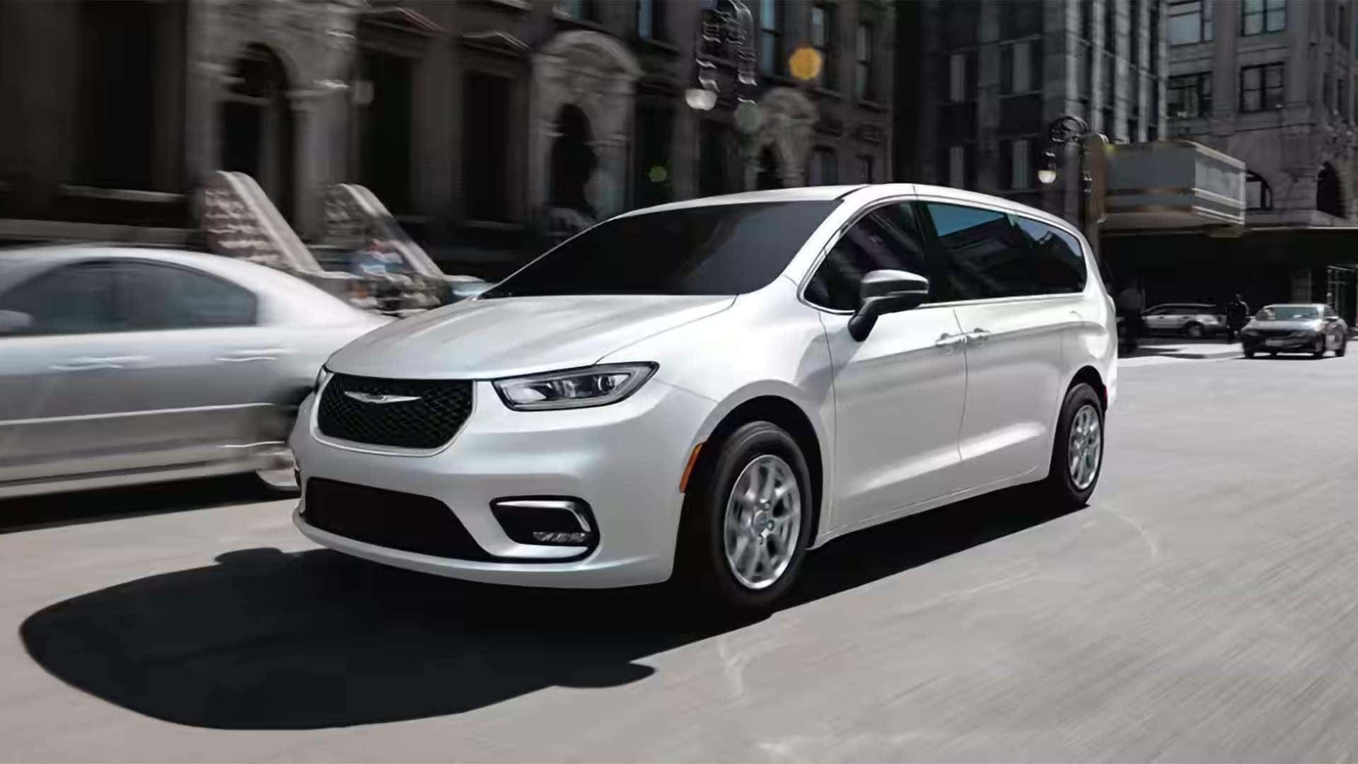 White 2026 Chrysler Pacifica driving through a city street showing exterior proportions and front grille design