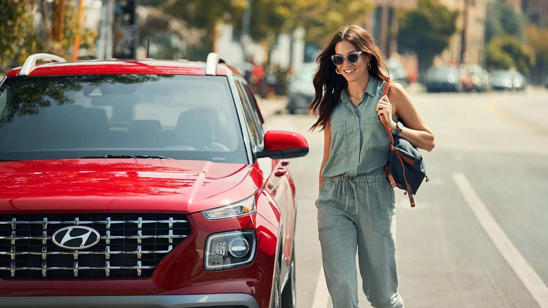Person walking beside a red 2026 Hyundai Venue on a city street, highlighting trim style, front grille, and urban practicality