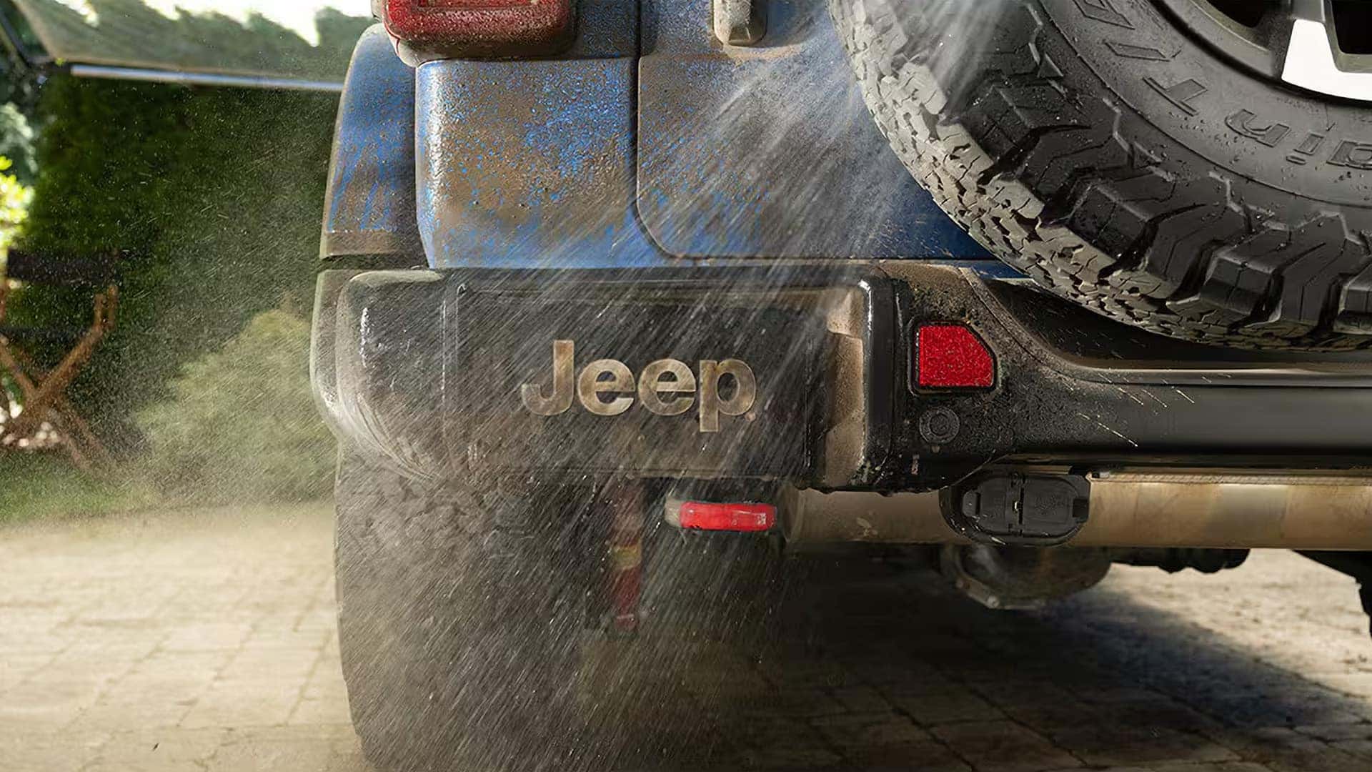 Rear close-up of a blue Jeep Wrangler showing spare tire, Jeep logo on mud flap, and water spray behind the bumper.