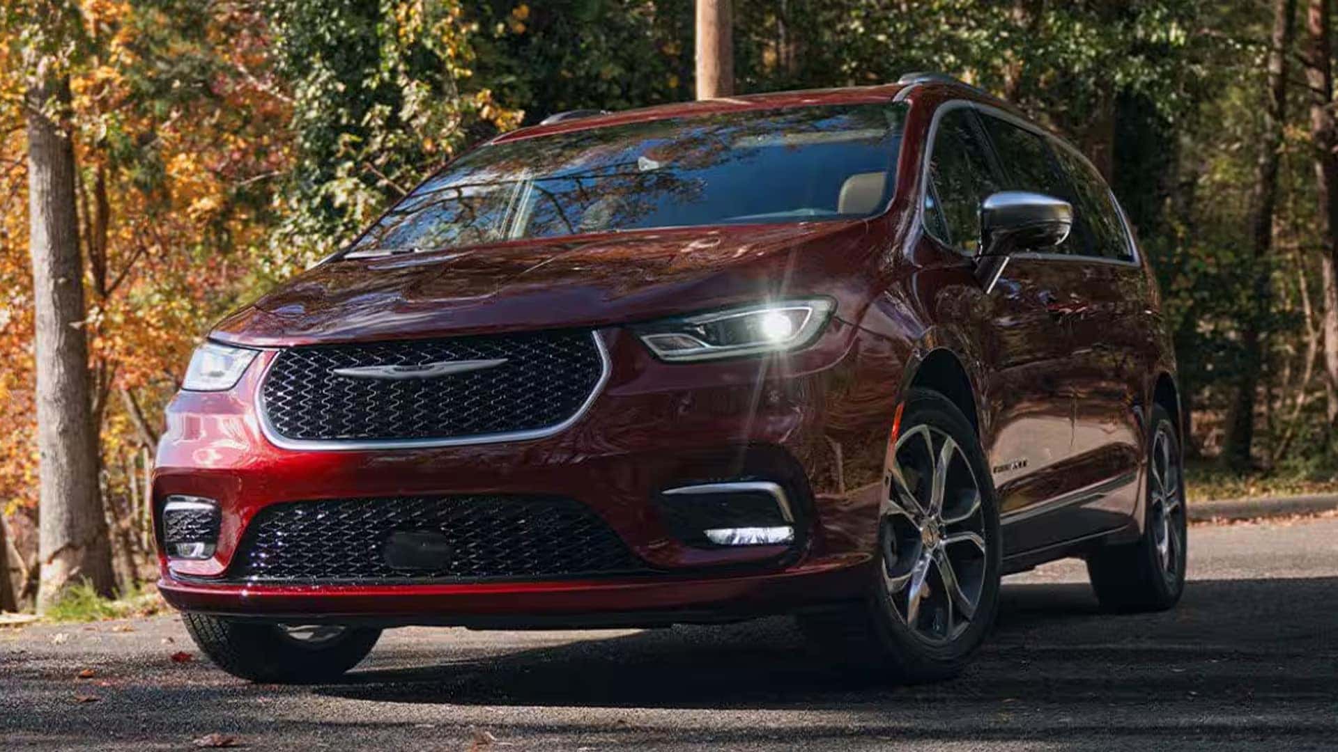 Red 2026 Chrysler Pacifica parked on a tree lined road, front three quarter view with alloy wheels and grille