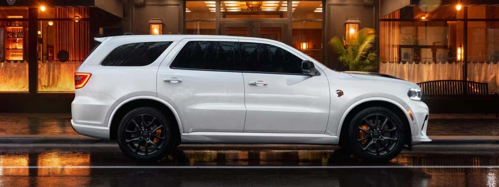 White 2026 Dodge Durango SRT Hellcat shown in side profile parked in front of a modern building at night, highlighting muscular SUV styling and performance stance