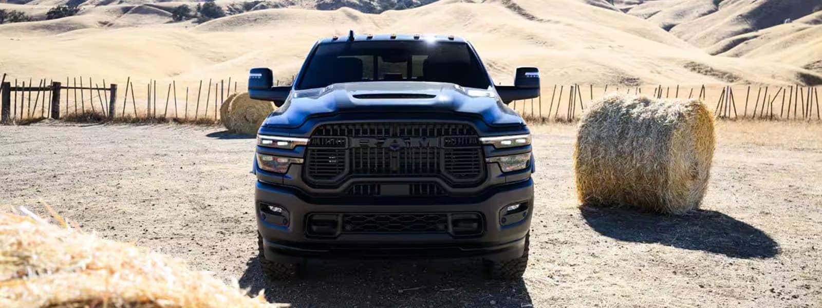 Front view of a 2026 RAM 2500 heavy-duty truck on a rural dirt lot with hay bales and hills behind it