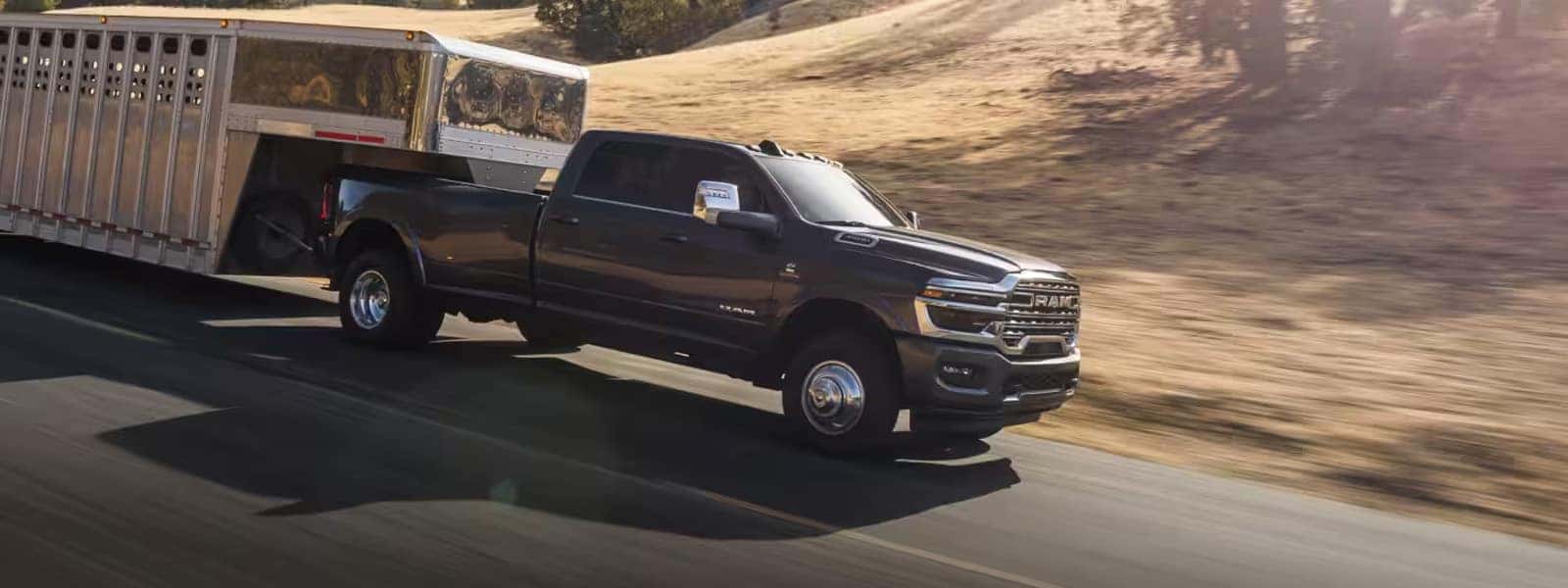 2026 RAM 3500 heavy duty pickup towing a large livestock trailer on a rural road, front three-quarter driving view