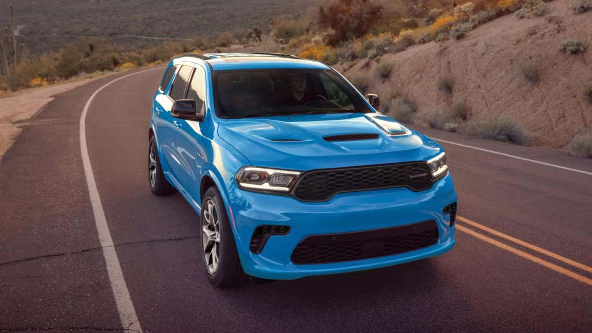 Bright blue 2026 Dodge Durango performance SUV driving on a winding desert road, front view highlighting aggressive grille and hood vents