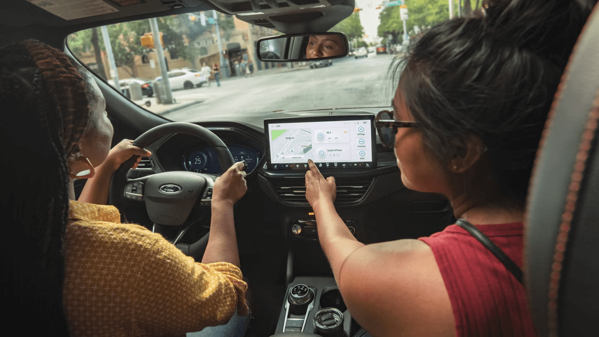 Rear-seat view of 2026 Ford Escape driving in city while passenger taps the center screen navigation