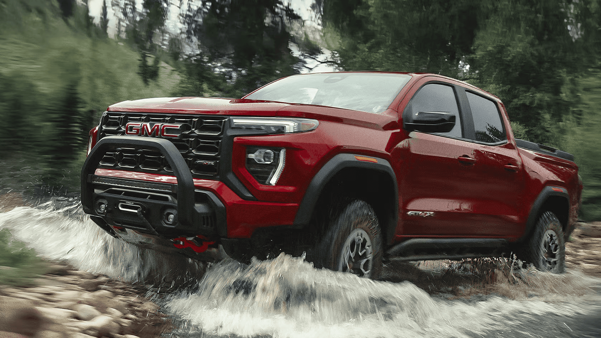 Red 2026 GMC Canyon AT4X splashing through rocky stream with off road tires and rugged front bumper.