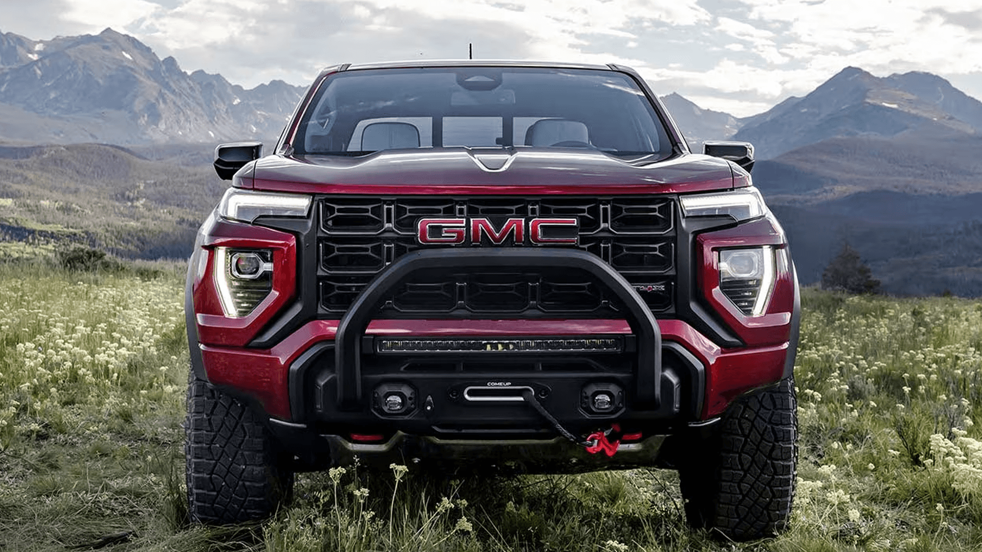 Front view of red 2026 GMC Canyon AT4X with off road bumper, winch, and LED lighting in mountain meadow.