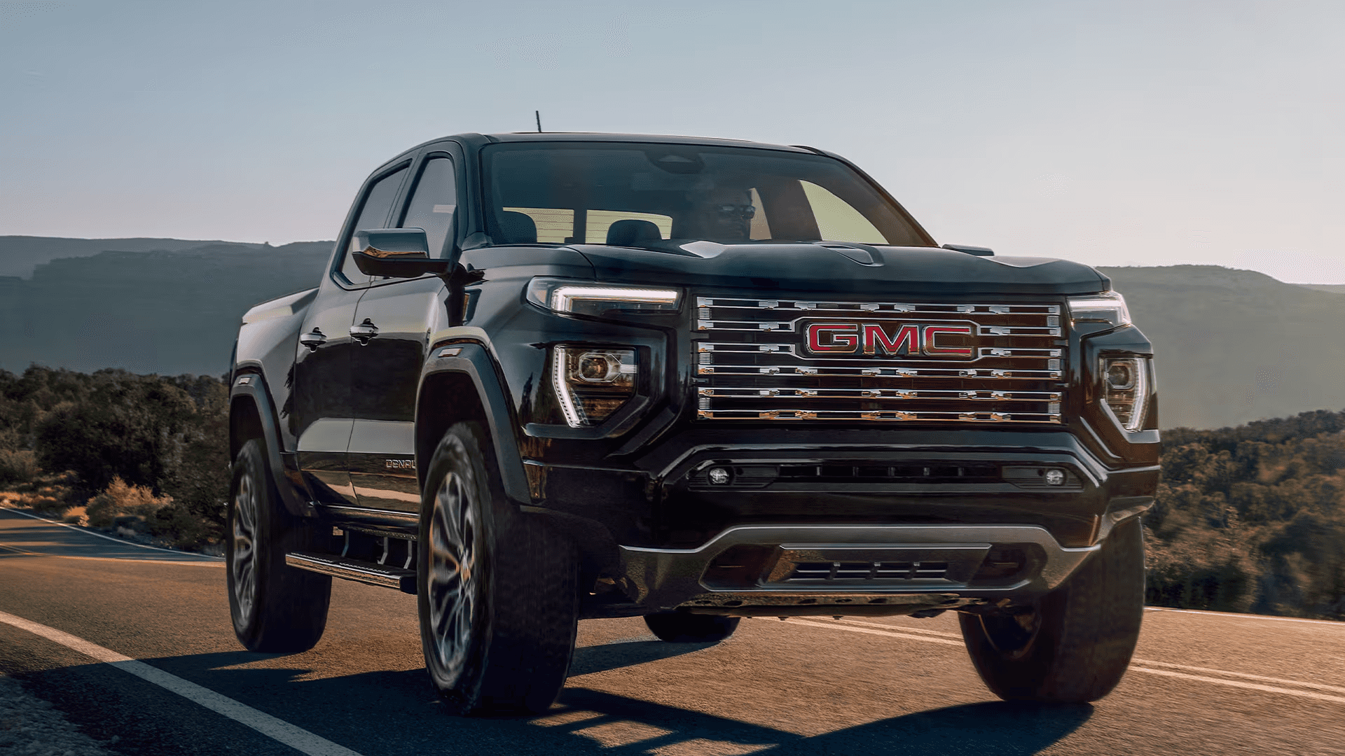 Black 2026 GMC Canyon Denali driving down desert road with mountains in the background.