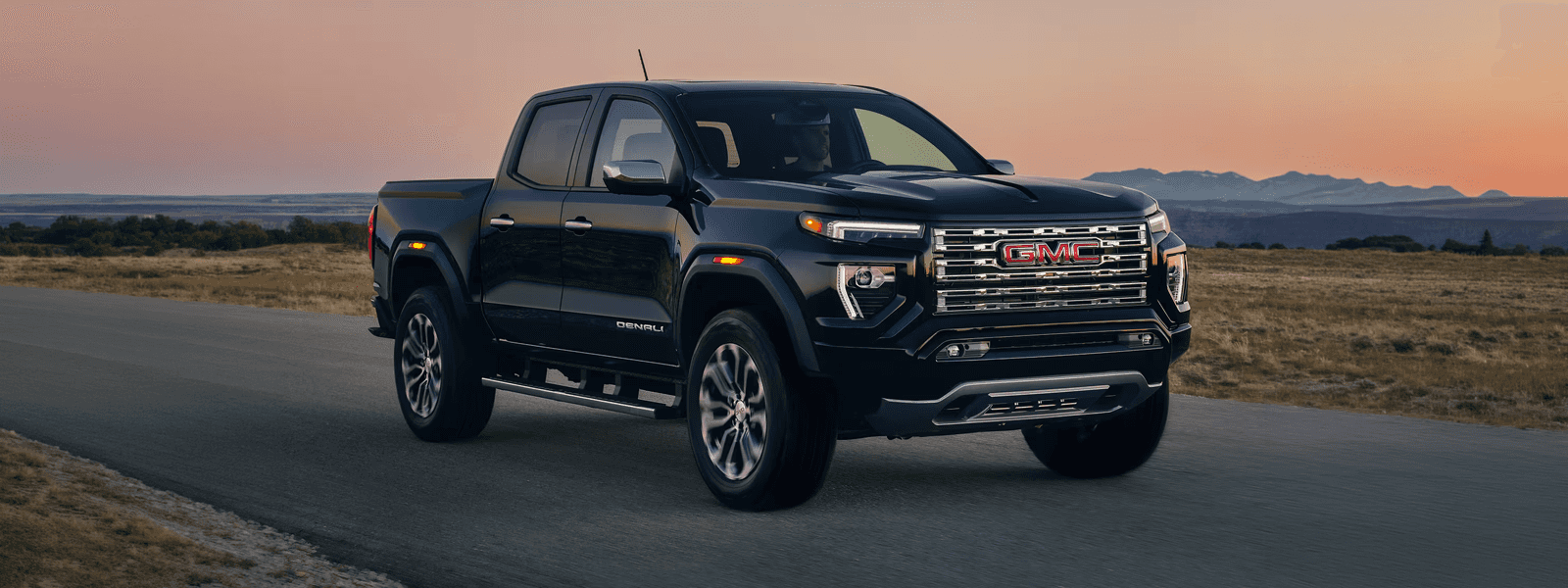Black 2026 GMC Canyon Denali driving on open highway at sunset with chrome grille and LED lighting.