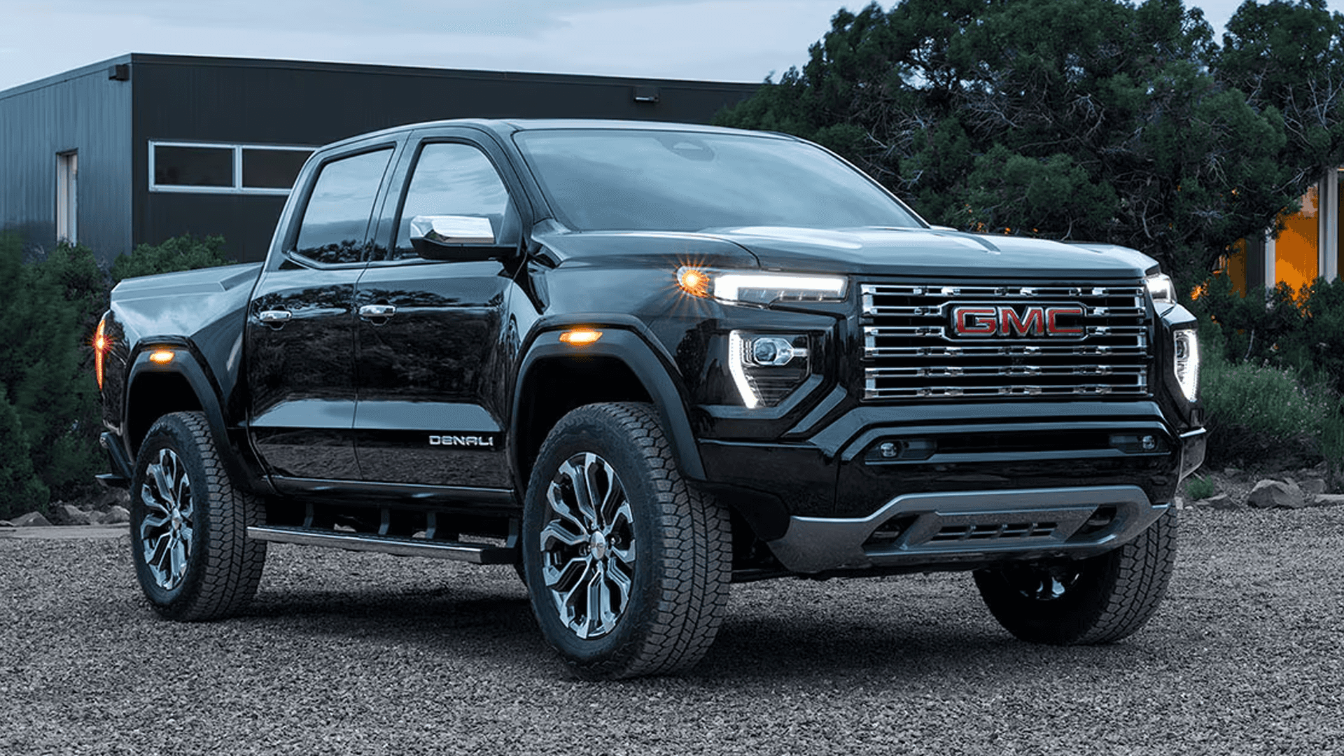 Black GMC Canyon Denali parked on gravel at dusk with LED headlamps and chrome grille illuminated