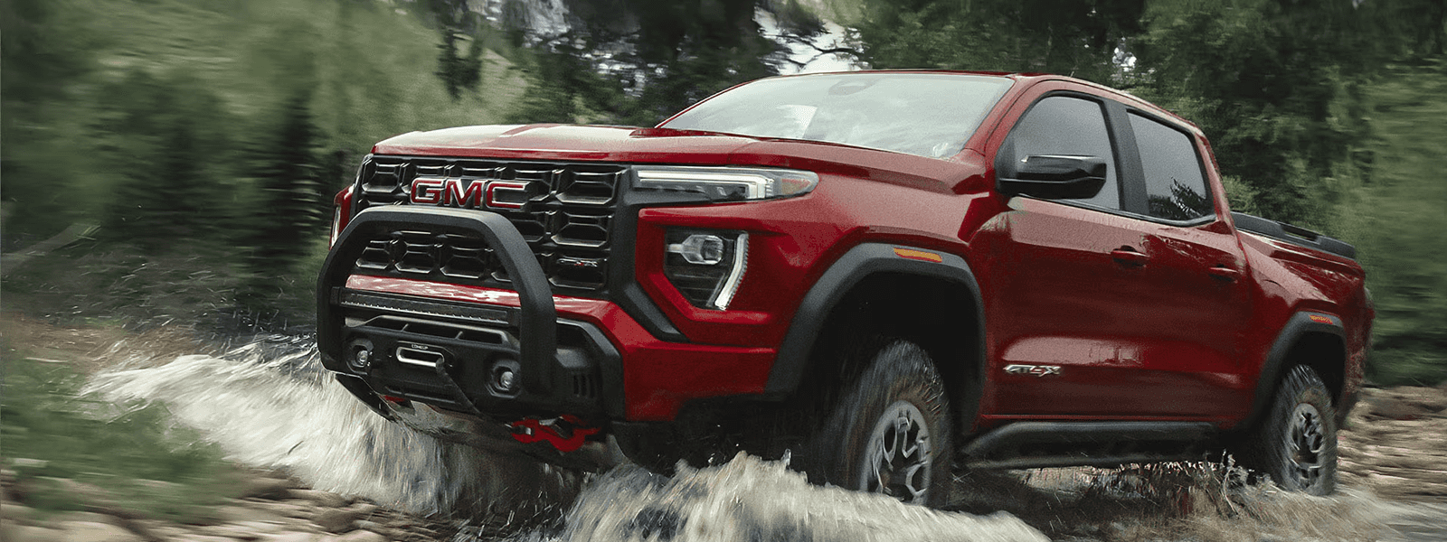 Red GMC Canyon AT4X splashing through shallow water on a forest trail, front three quarter action view