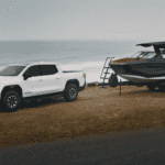 White GMC Sierra EV parked beside a large boat on a trailer near the ocean, front three-quarter view