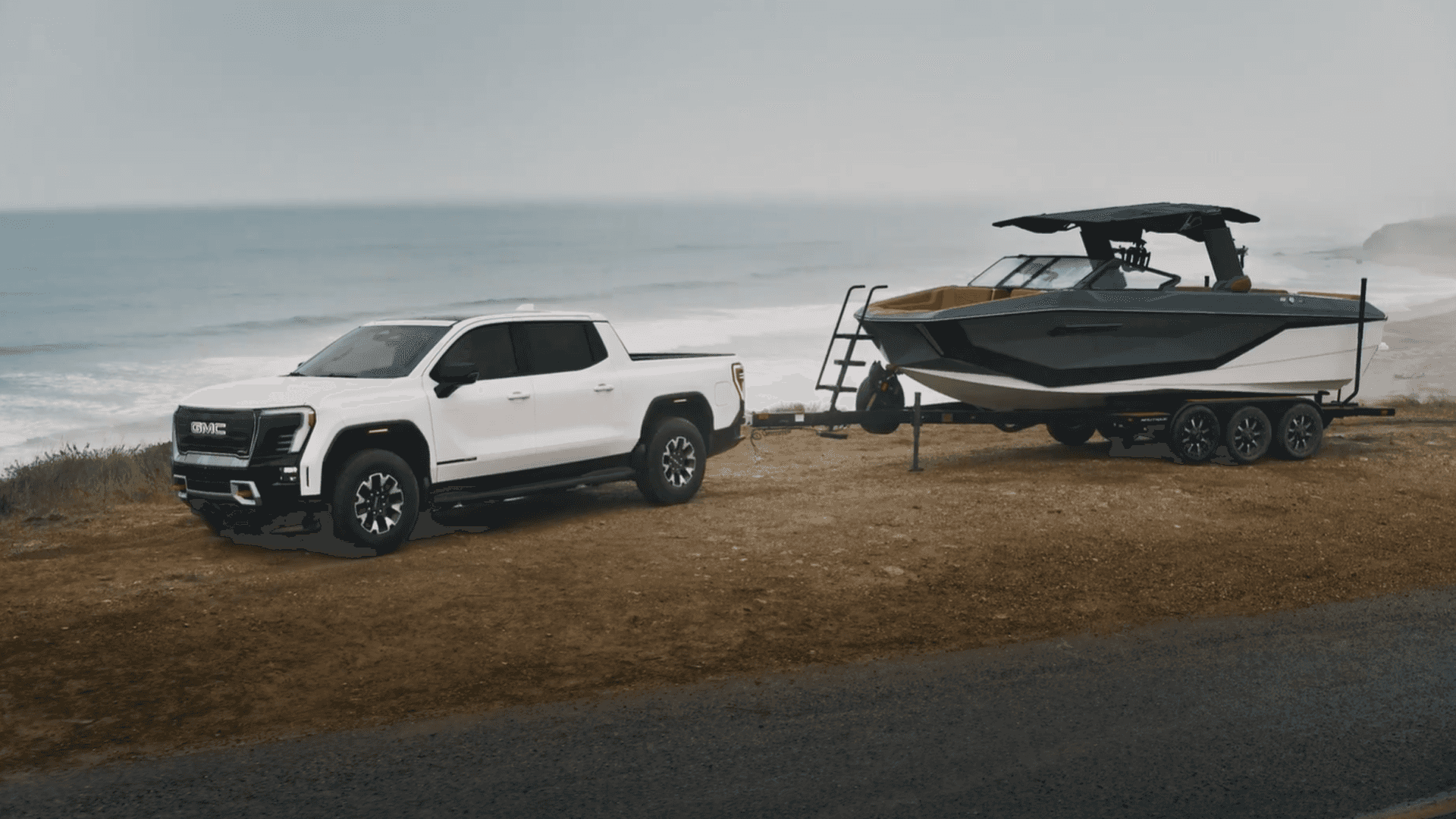White GMC Sierra EV parked beside a large boat on a trailer near the ocean, front three-quarter view