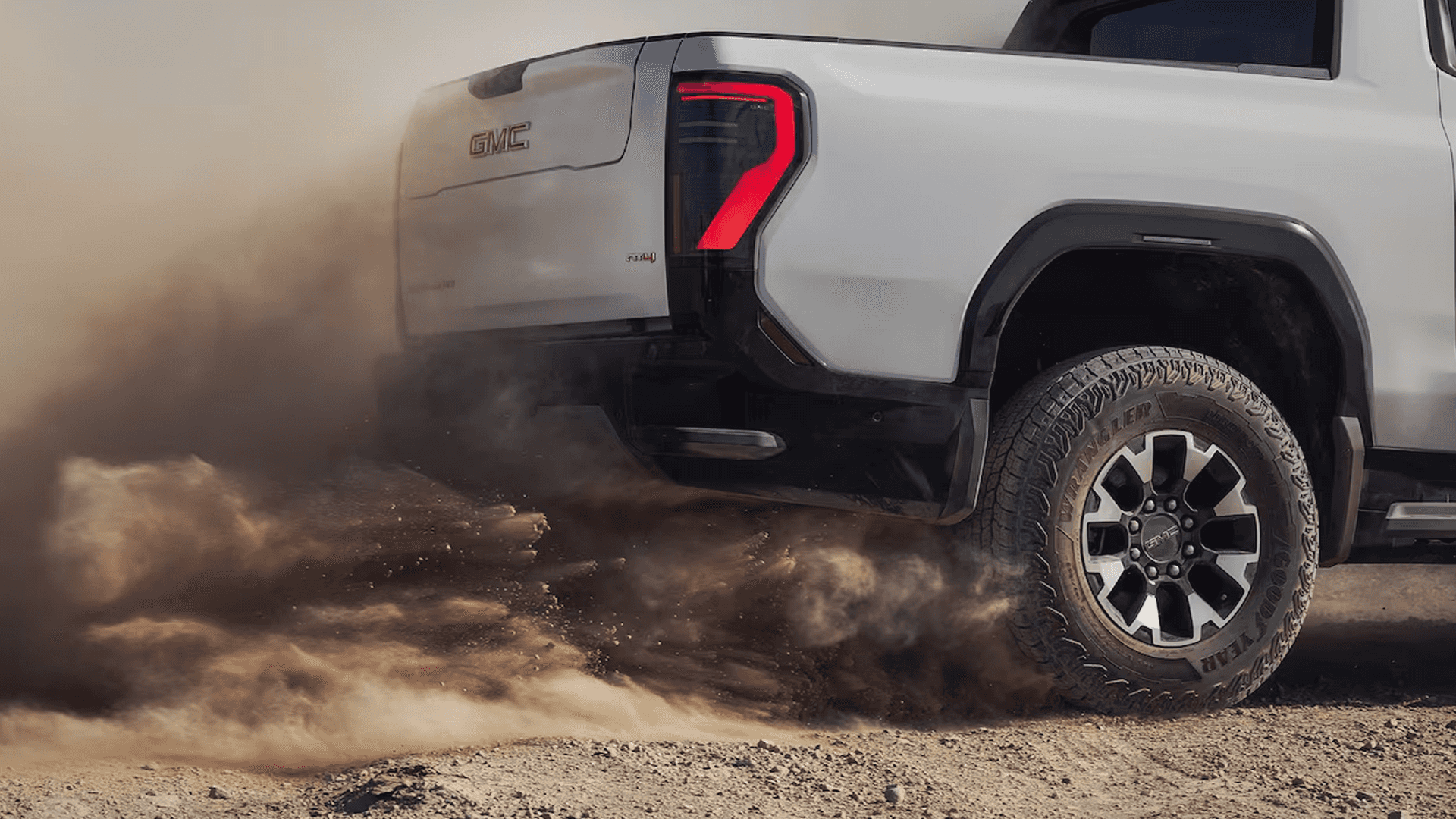 Close view of a white GMC truck's rear wheel and taillight kicking up dust on a dirt surface