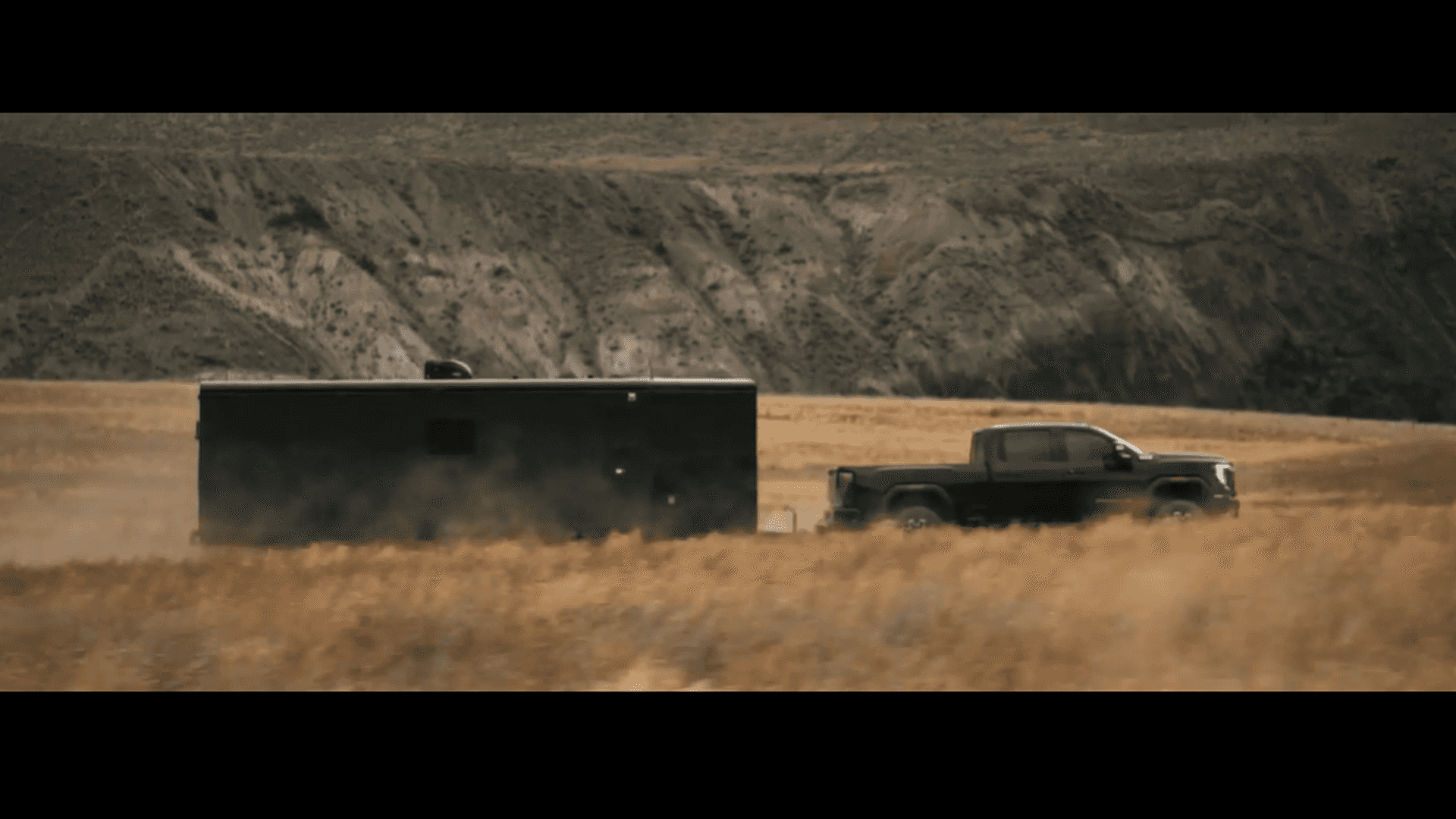 Dark GMC pickup towing a large enclosed trailer across a dry field with rocky hills in the background