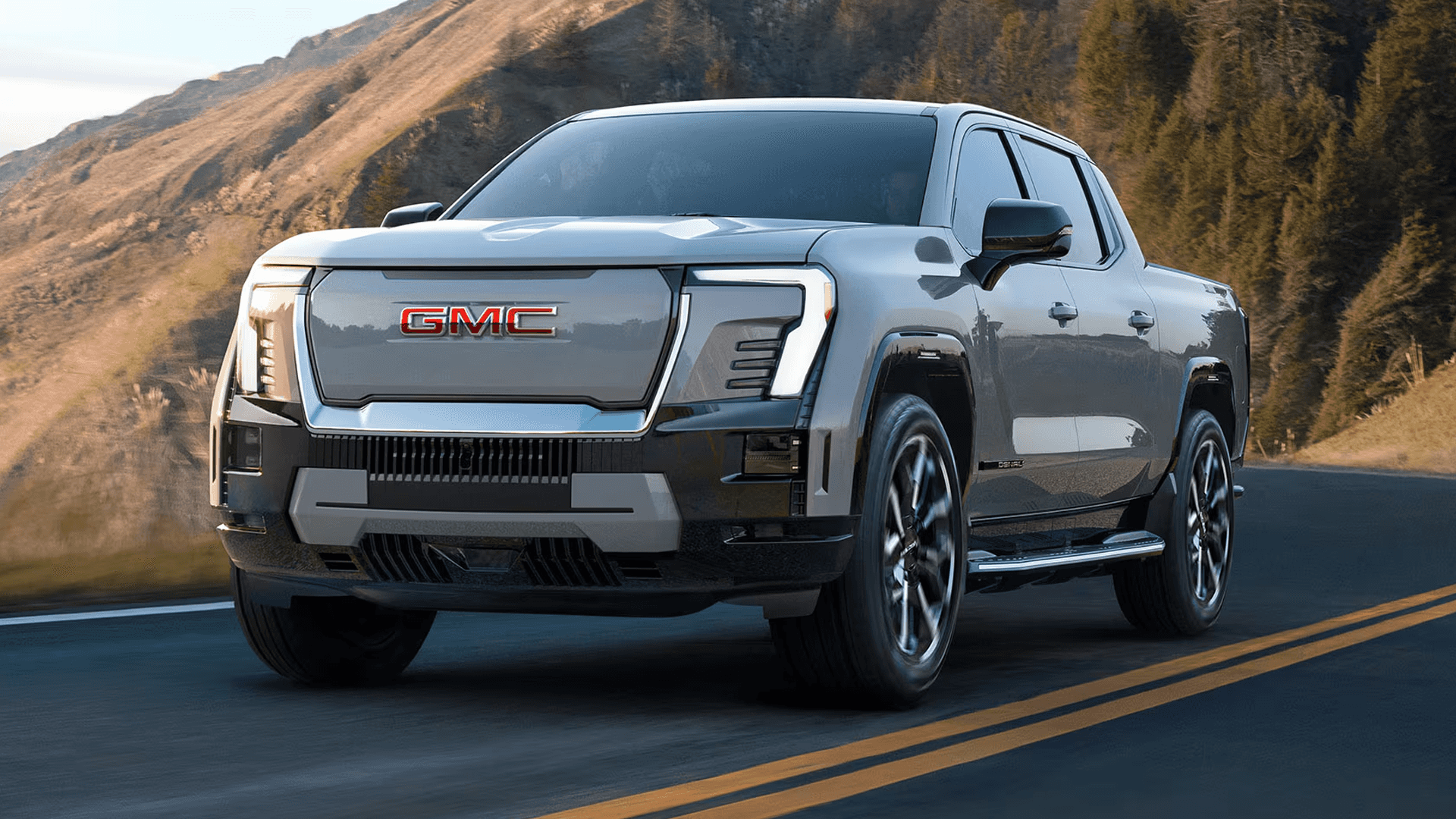 Gray GMC Sierra EV driving on a mountain road, front three-quarter view with forested hills in the background