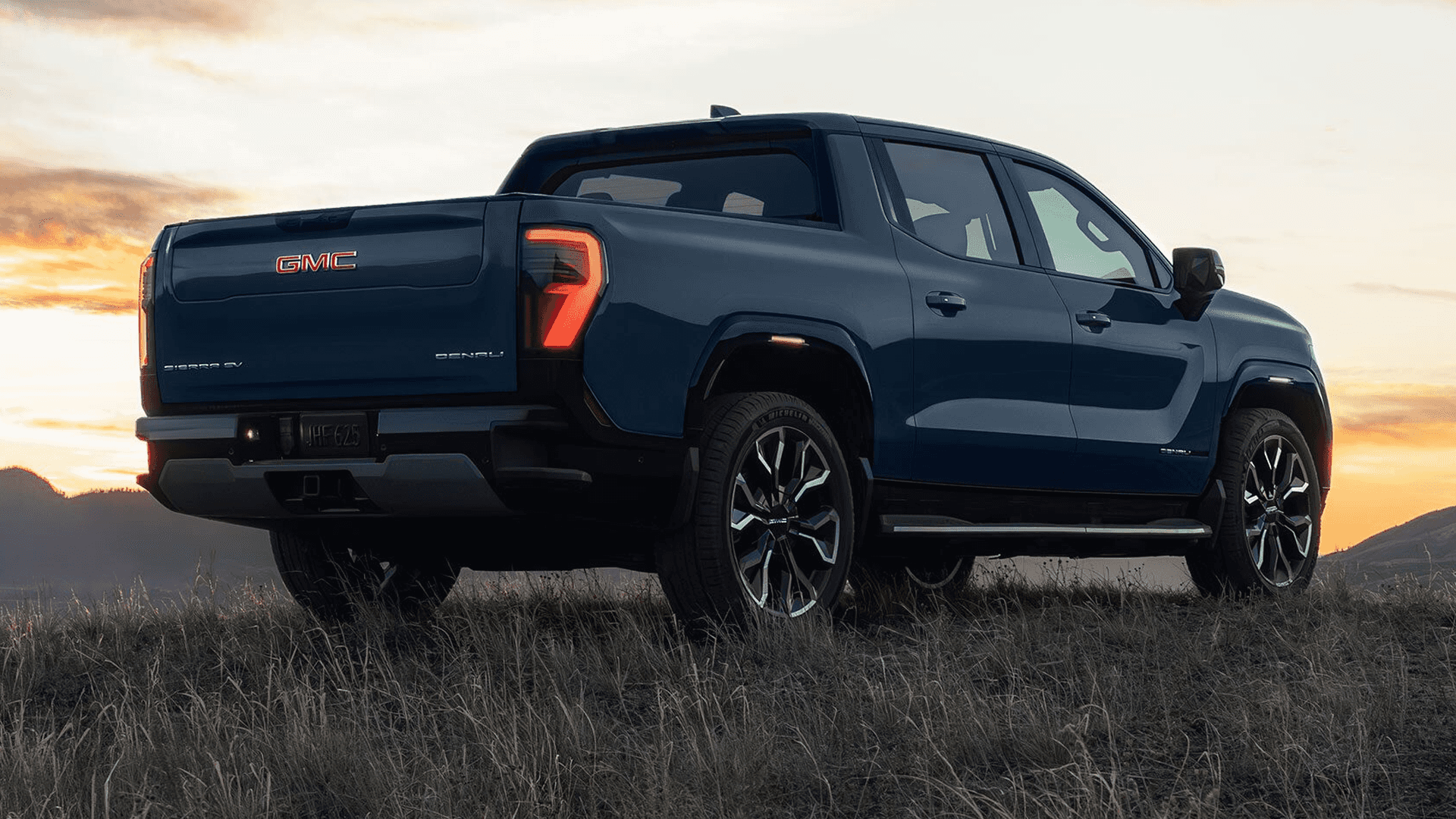 Blue GMC Sierra EV parked in a grassy field at sunset, rear three-quarter view with illuminated rear GMC badge