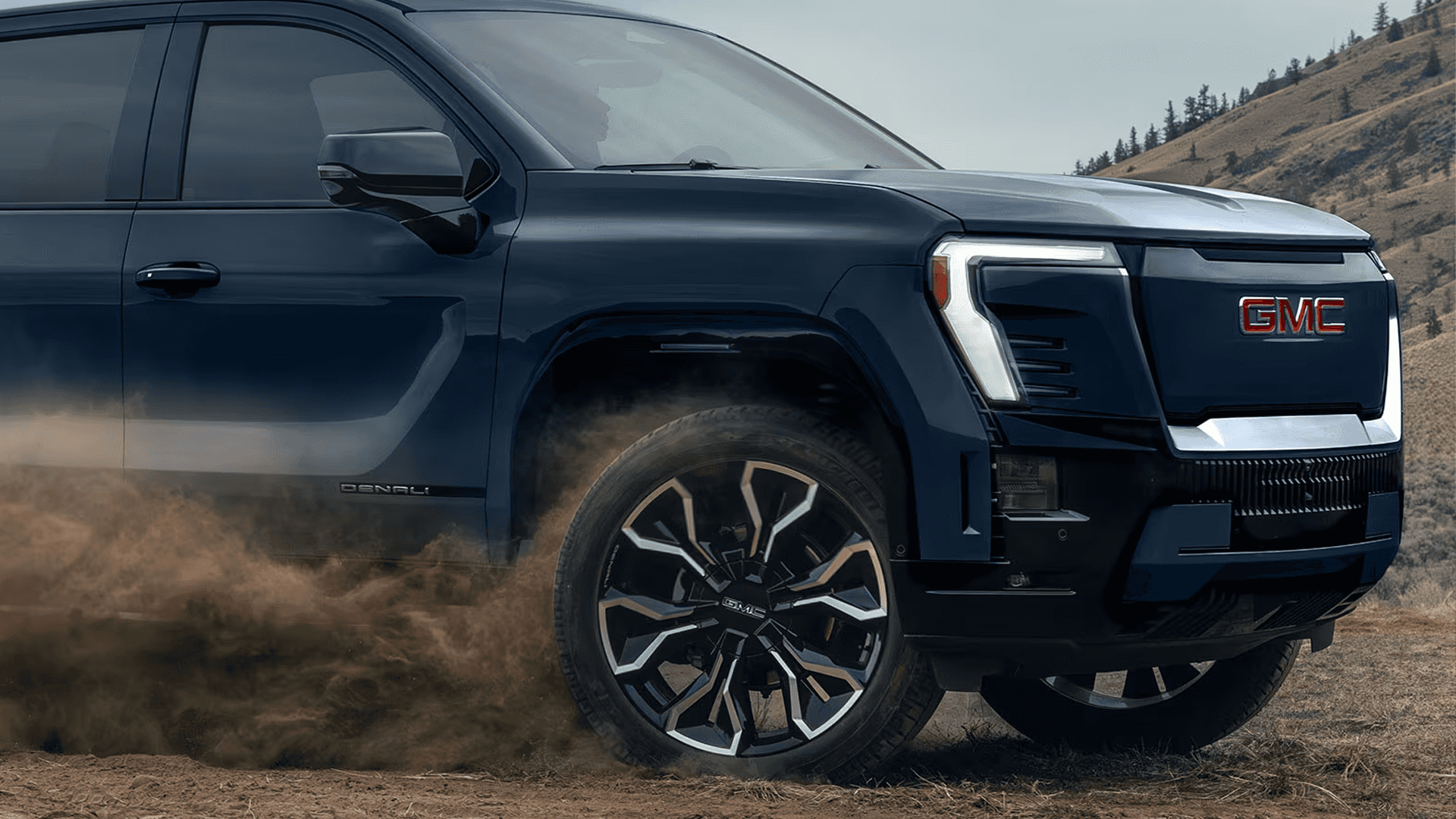 Front side view of a blue GMC Sierra EV kicking up dust, showing LED lighting, grille, and large alloy wheel