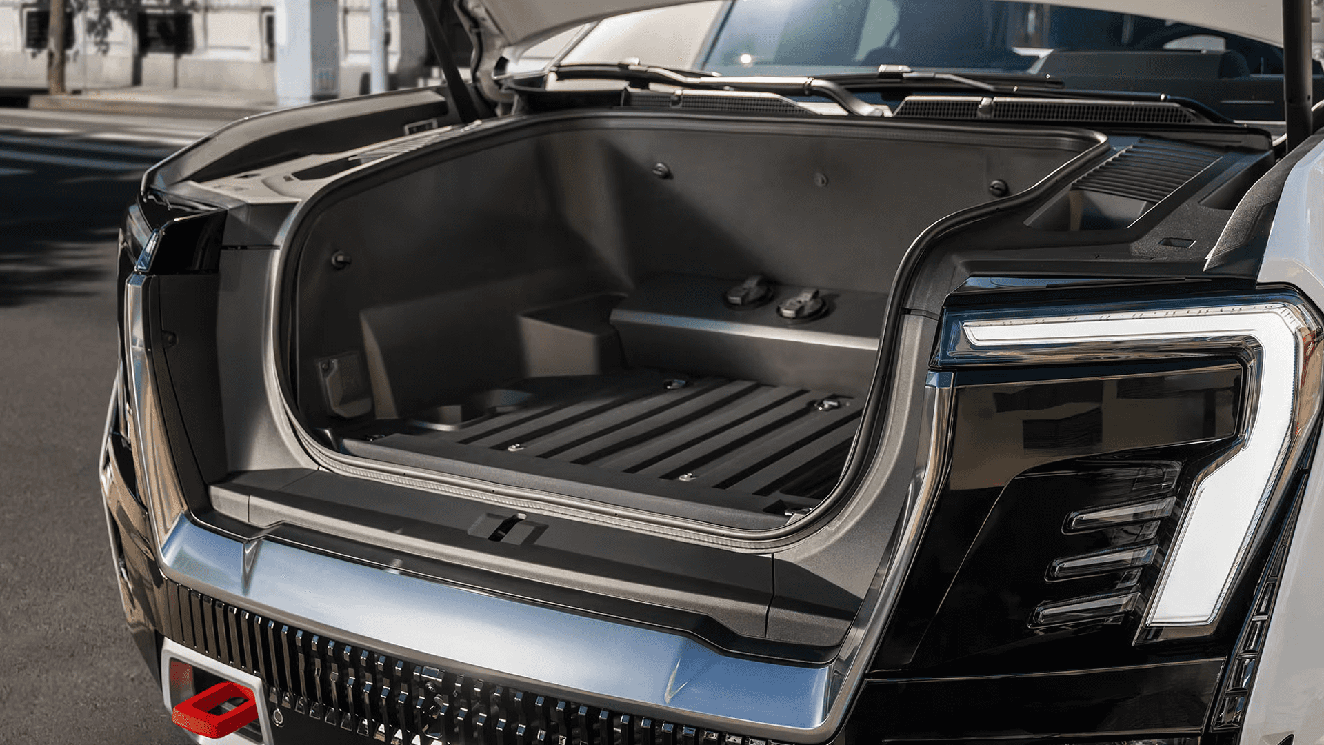 Open front trunk on a GMC Sierra EV showing the storage compartment, front light signature, and lower red tow hook