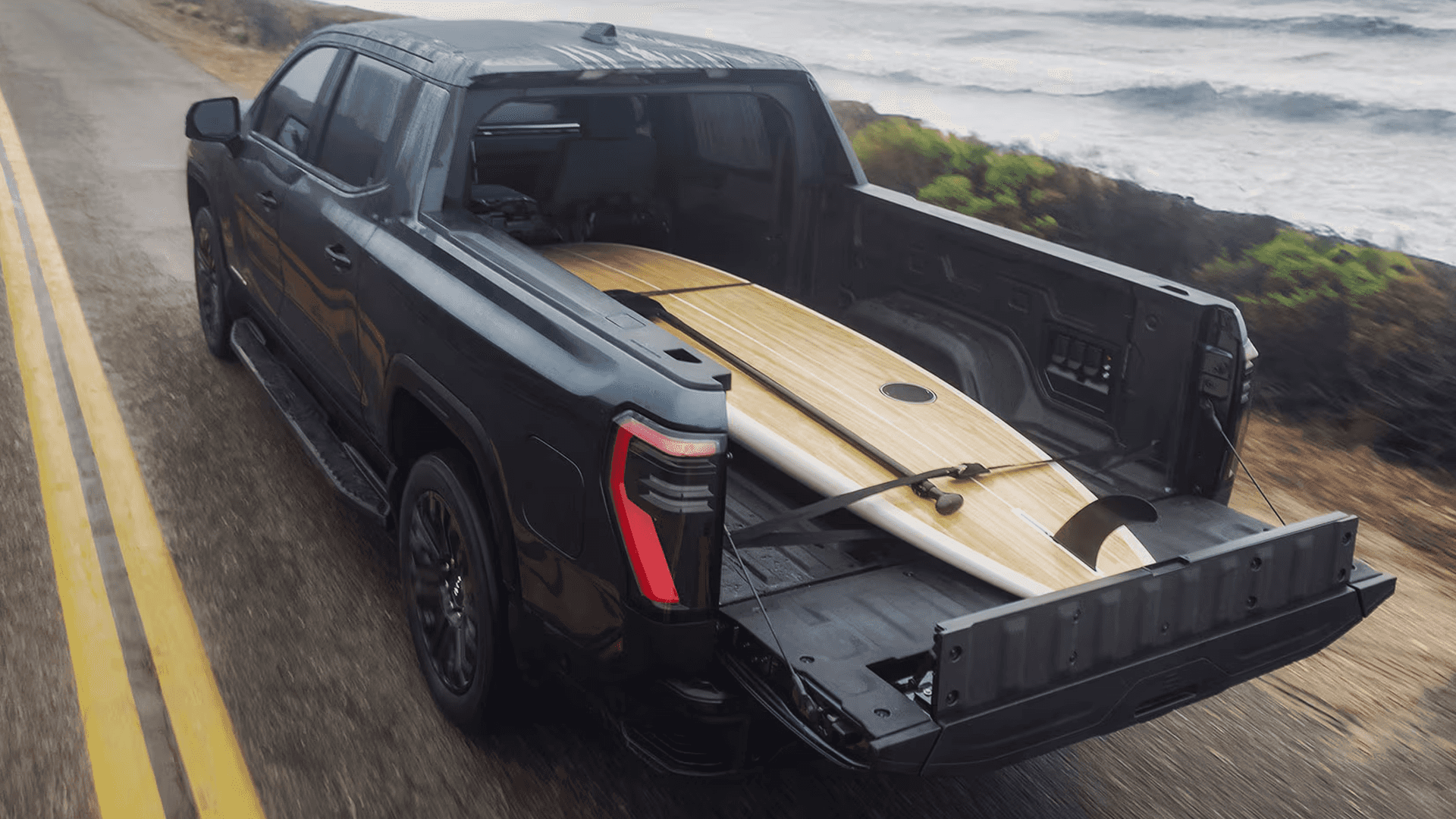 GMC Sierra EV driving beside the coast with the tailgate down and a surfboard secured in the truck bed