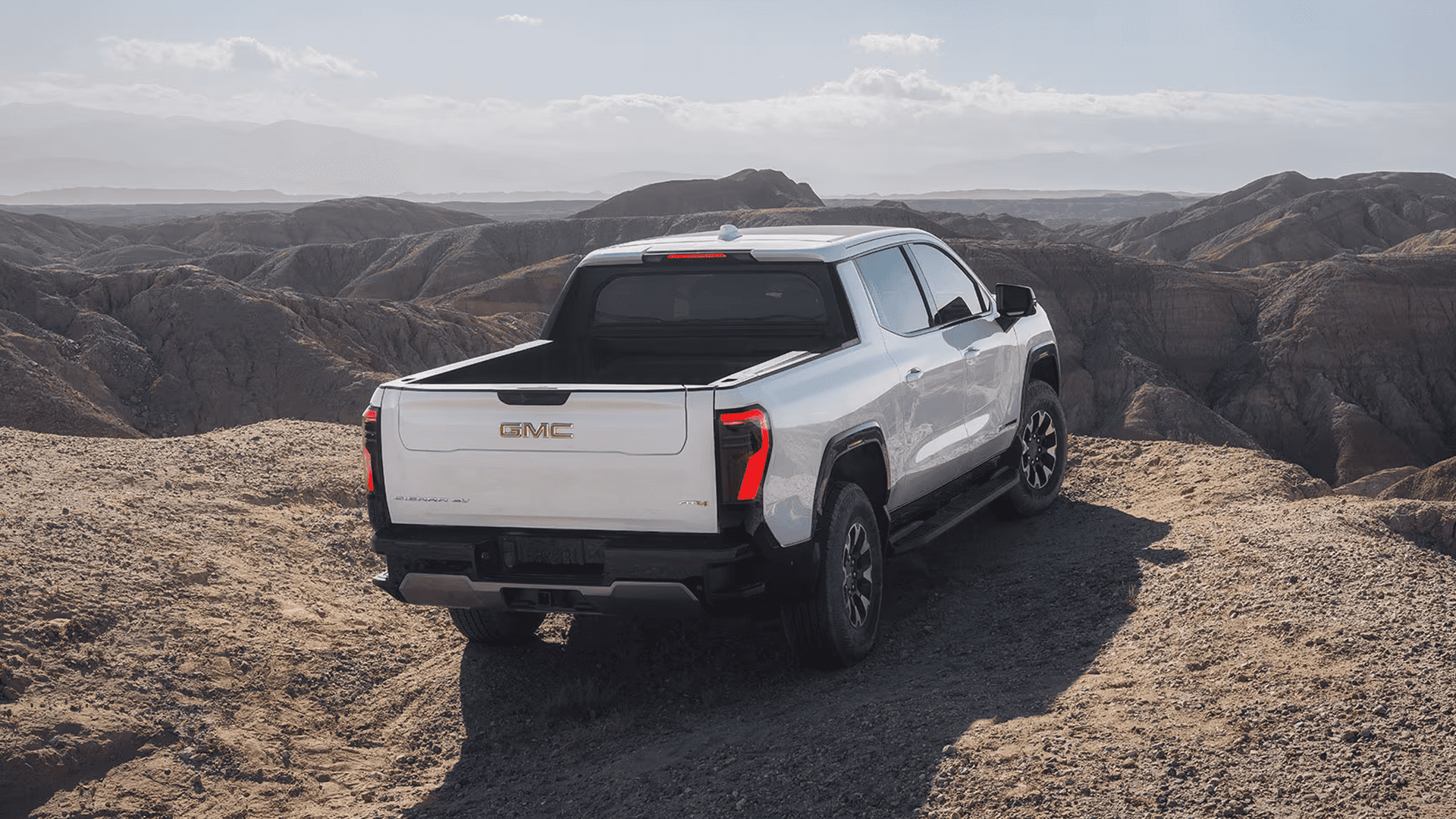 White GMC Sierra EV parked on rocky desert terrain, rear three-quarter view with canyon landscape in the background