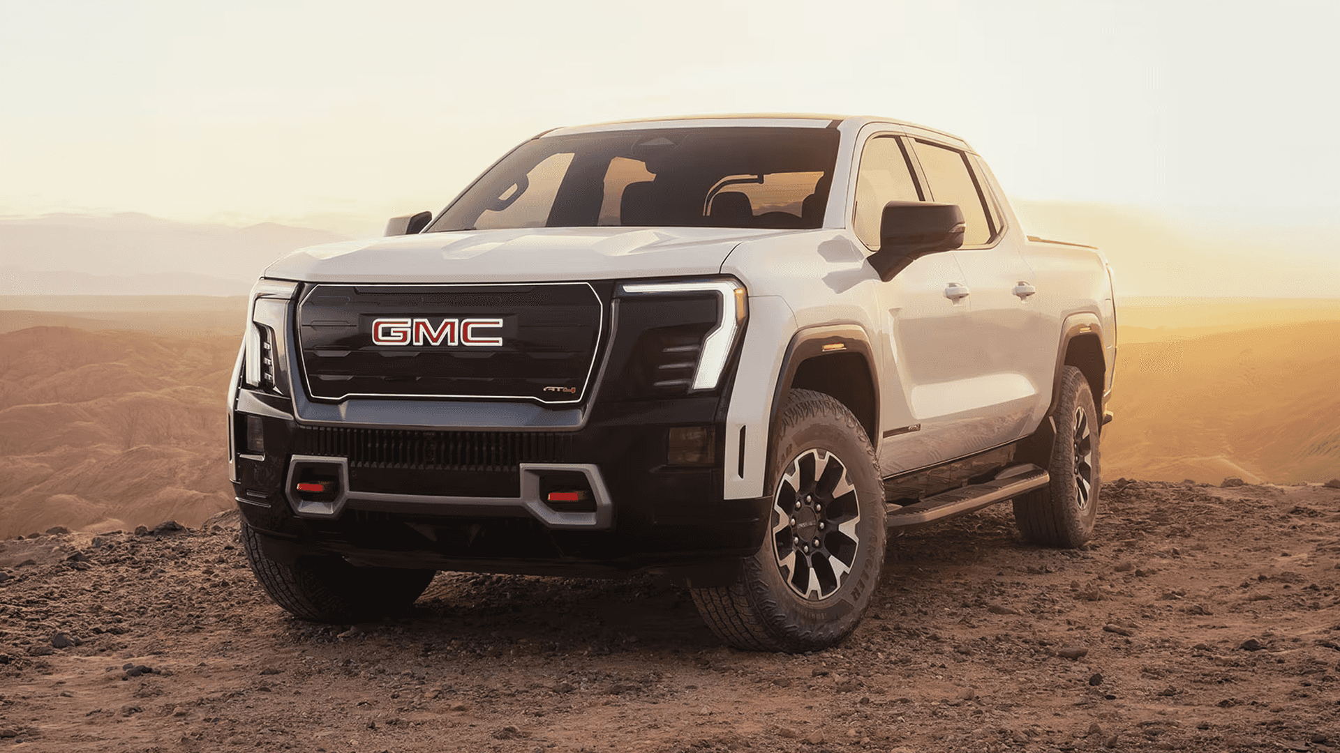 White GMC Sierra EV parked on rocky terrain at sunset with mountains in the distance