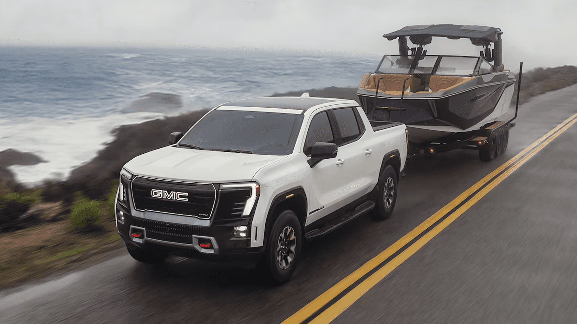 White GMC Sierra EV towing a large boat on a trailer along a coastal road beside the ocean