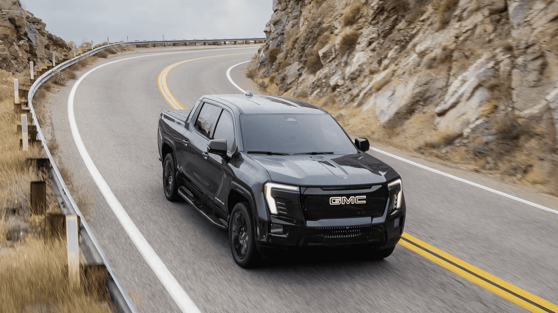 Dark GMC Sierra EV driving on a winding mountain road bordered by rock walls and guardrails
