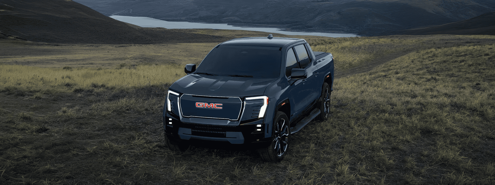 Blue GMC Sierra EV parked on grassy terrain with a lake and rolling hills in the background