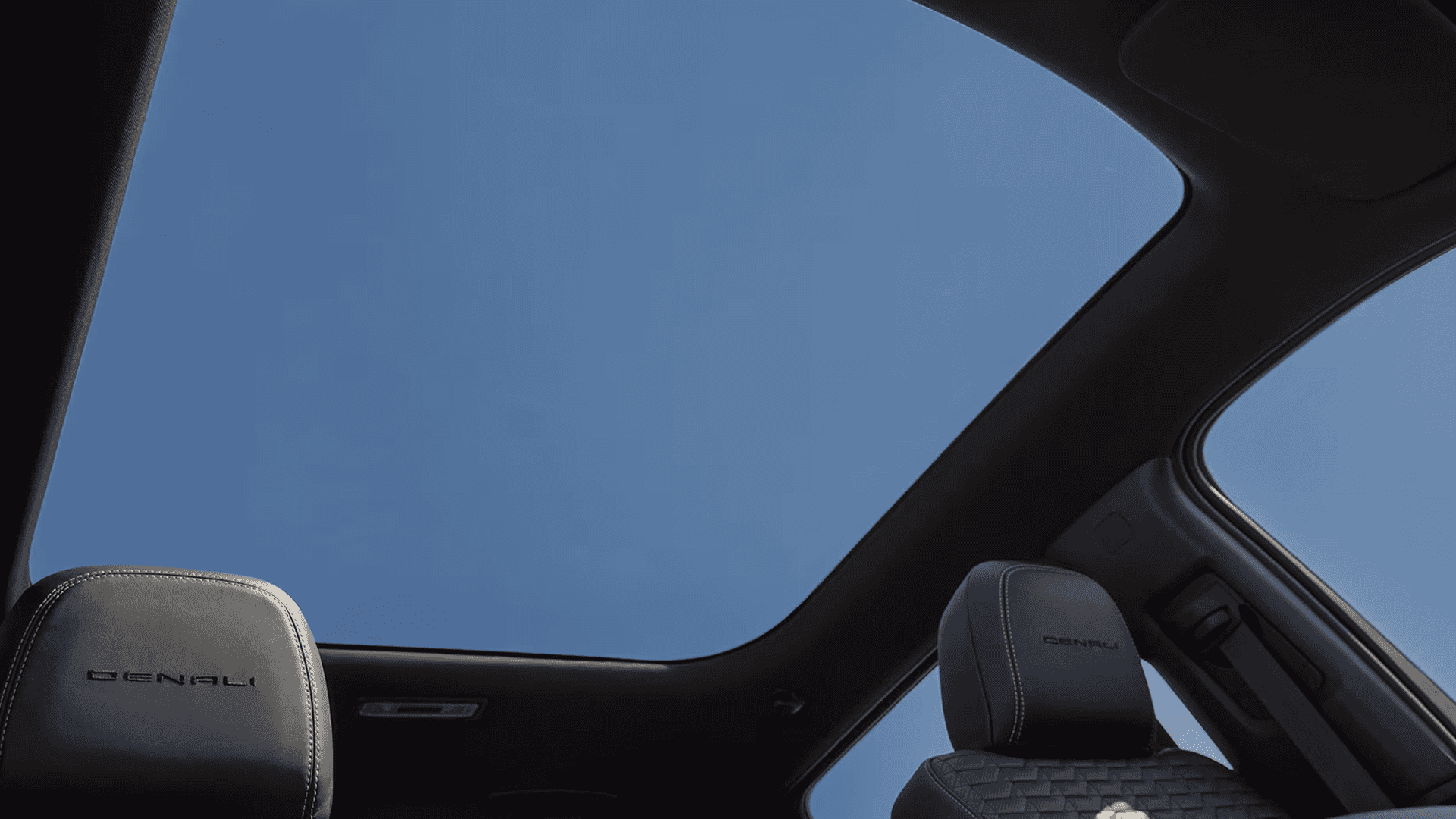 View through panoramic glass roof with blue sky and two Denali seat headrests visible below