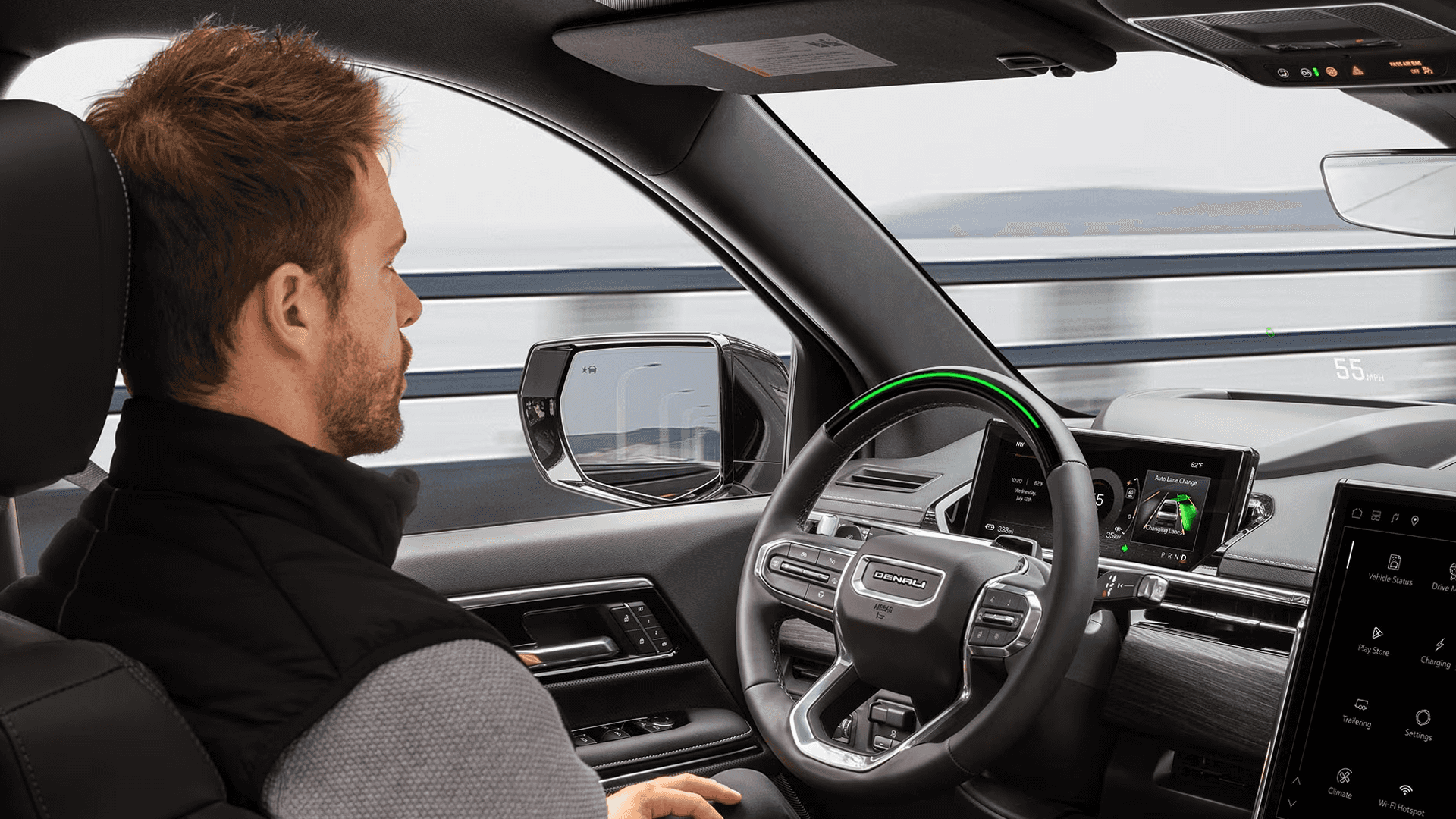 Driver seated in GMC Sierra EV cabin with green steering wheel light and highway barrier visible outside