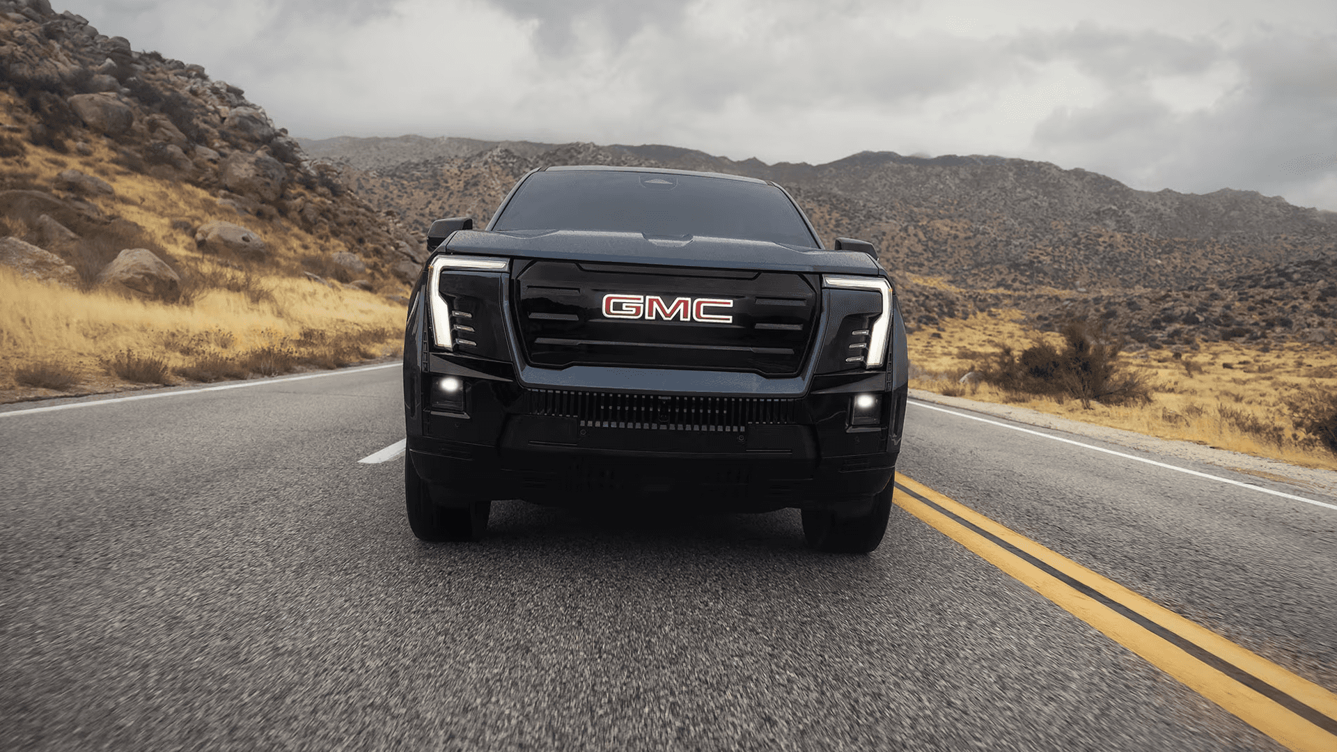 Front view of a black GMC Sierra EV driving on a two-lane road with rocky hills and cloudy sky behind