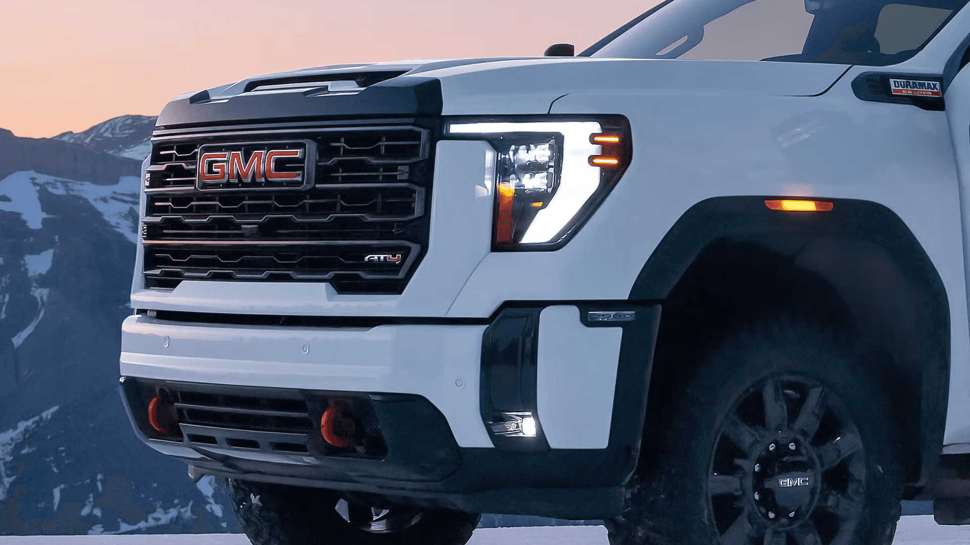 Close view of a white GMC Sierra HD AT4 front end with LED signature lighting and grille at dusk in the mountains.