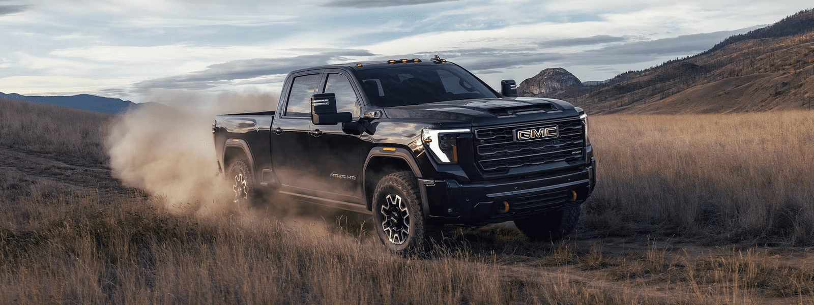 Black GMC Sierra HD AT4X driving through tall grass with dust behind it and mountains in the background.