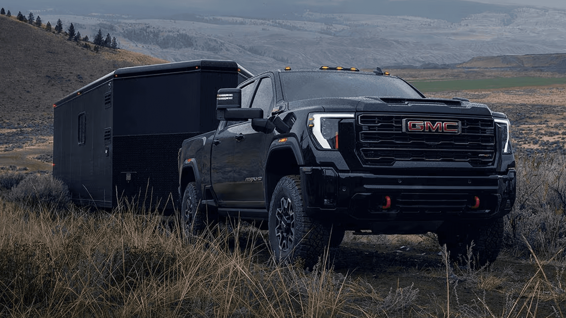 Black GMC Sierra HD AT4X towing a large enclosed trailer through grassy hills with red recovery hooks visible