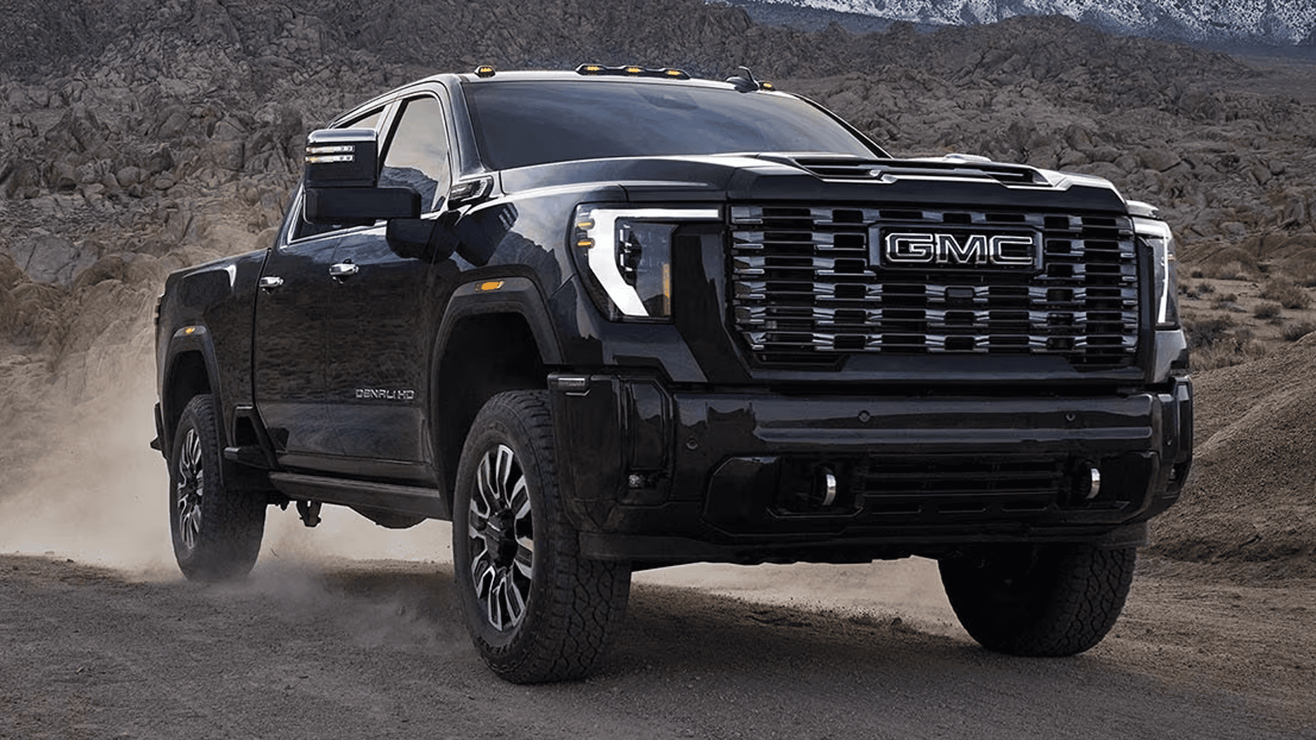 Black GMC Sierra HD Denali Ultimate driving on a dusty mountain road with large grille and tow mirrors visible