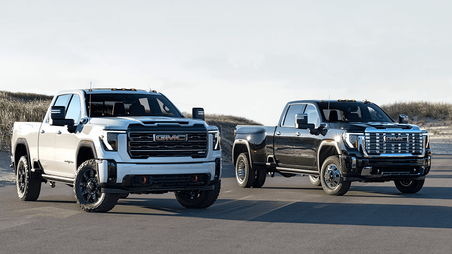 White GMC Sierra HD AT4 and black Sierra HD Denali parked side by side on pavement near coastal dunes