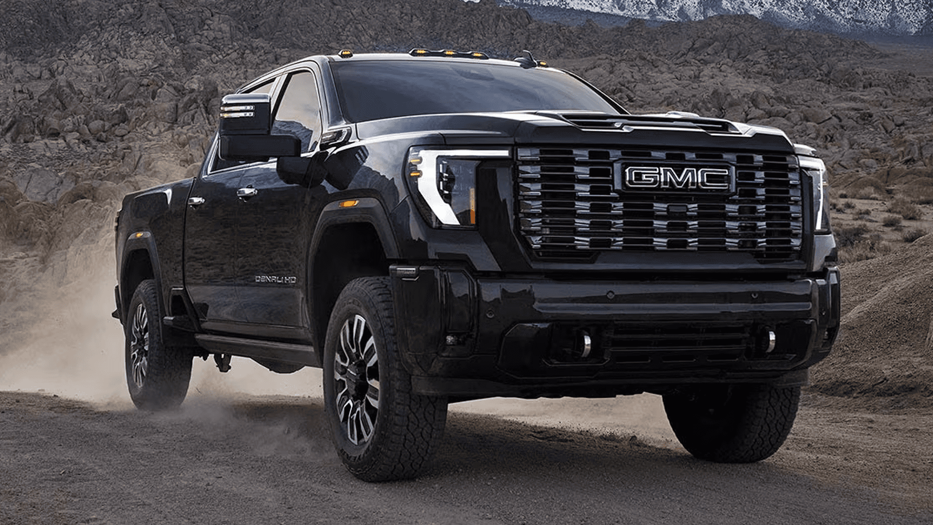Front three quarter view of a 2026 GMC Sierra HD Denali driving on a rugged desert road with mountains in the background.