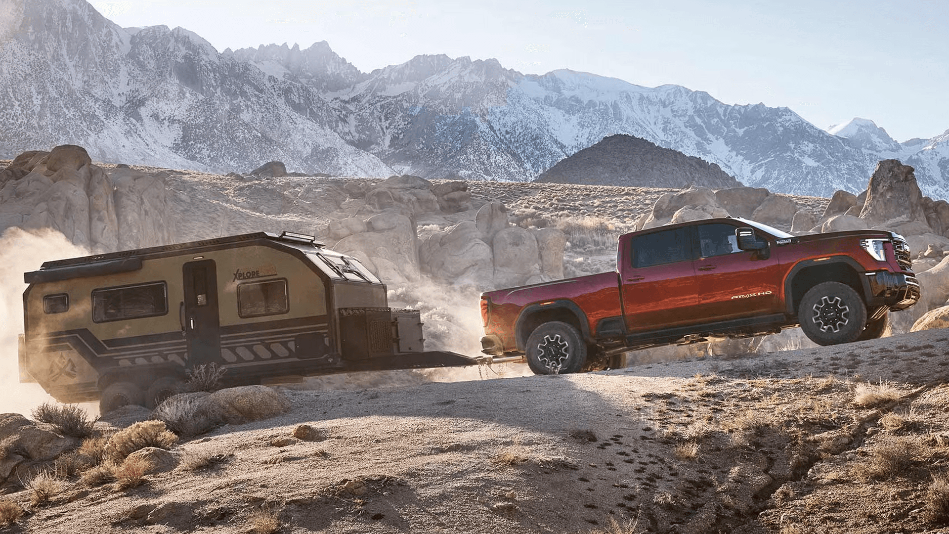 Red 2026 GMC Sierra HD towing a black travel trailer uphill through rocky desert terrain below snowy mountains.