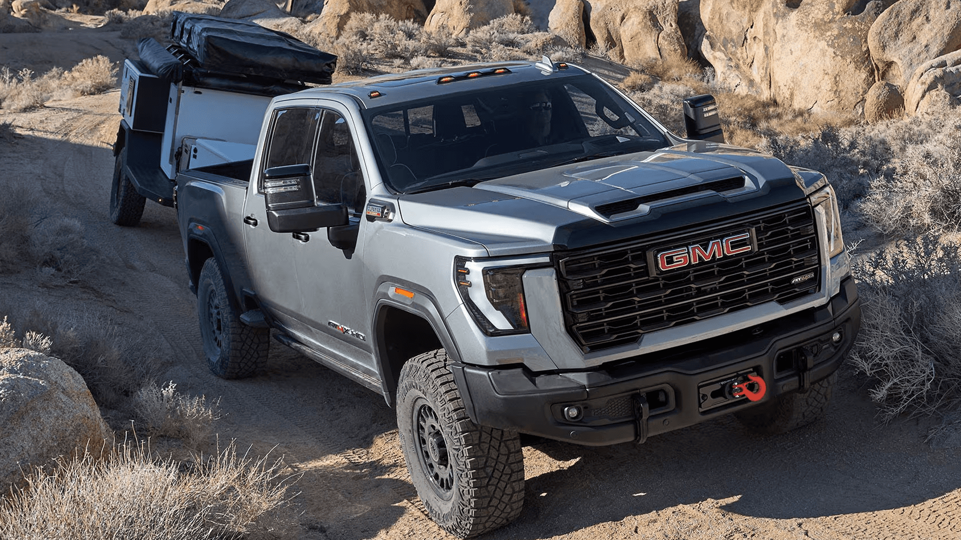 Gray 2026 GMC Sierra HD AT4X towing an overland camper through rocky desert terrain with extended tow mirrors.