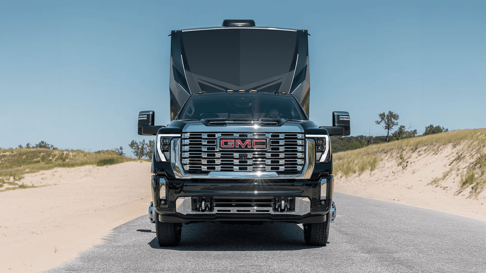 Front view of a black 2026 GMC Sierra HD towing a large RV trailer on a paved road between sandy dunes.
