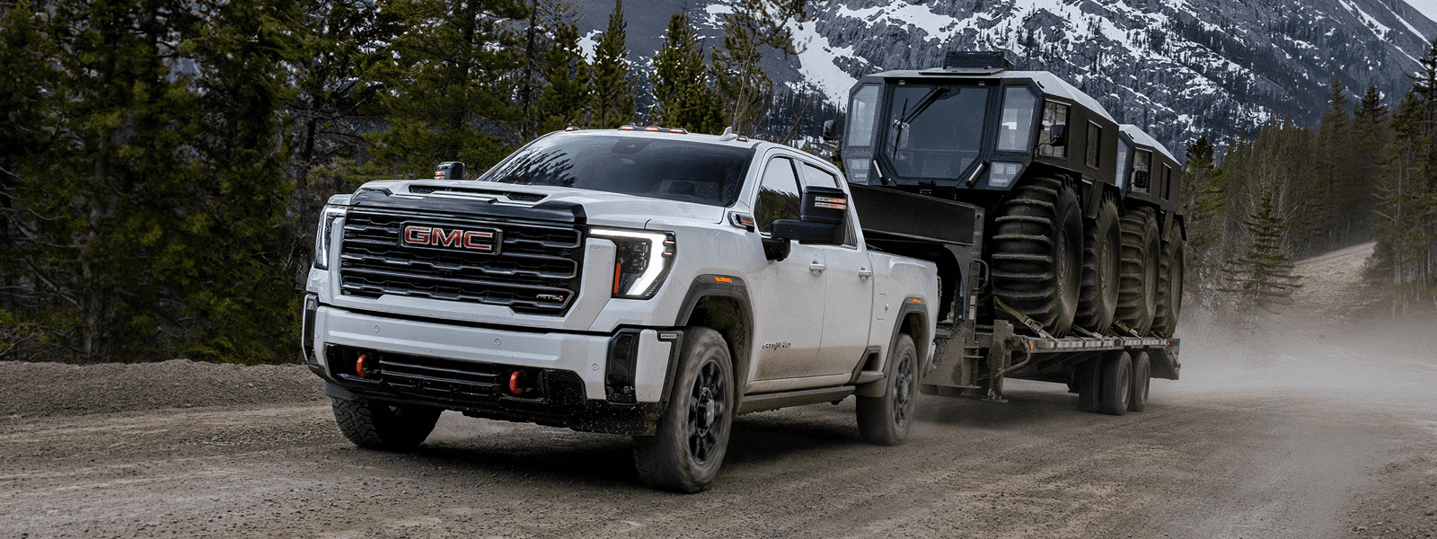 White 2026 GMC Sierra HD AT4X towing a huge off-road trailer on a dirt road with mountains in the background.