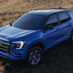 Blue 2026 GMC Terrain Denali parked on grassy hillside showing bold grille, C shaped LED headlights, and sculpted body design.