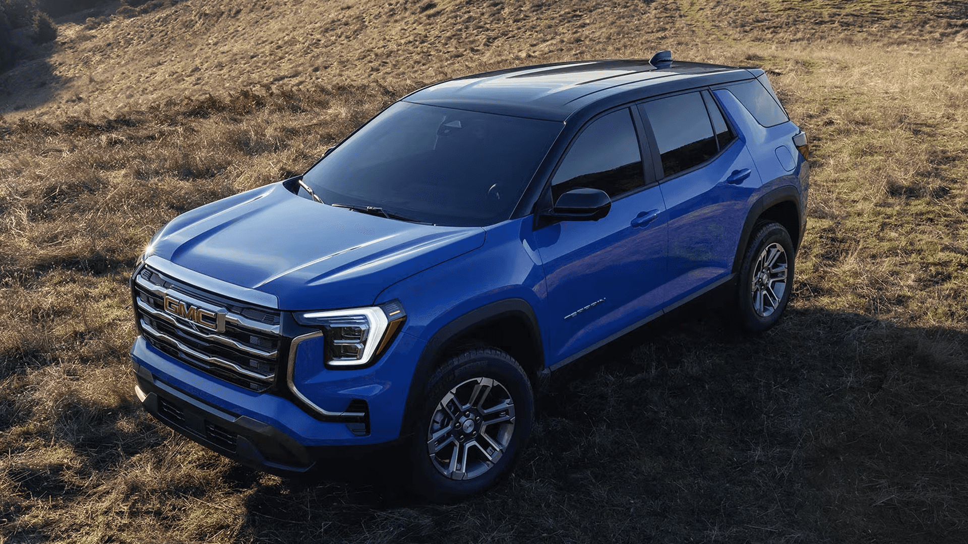 Blue 2026 GMC Terrain Denali parked on grassy hillside showing bold grille, C shaped LED headlights, and sculpted body design.